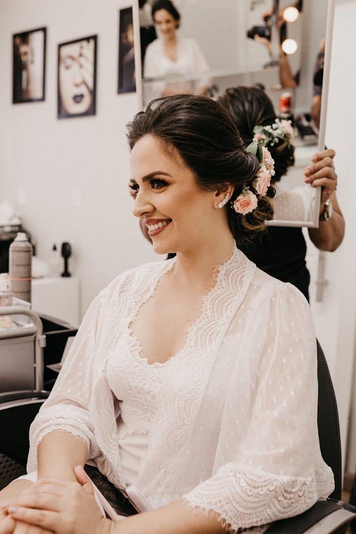 Bride smiling in salon while hairstylist prepares elegant hairstyle with flowers.